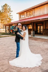 Aron and Kaitlin stand close together, smiling in front of the weathered wooden barn at Whispering Pines, surrounded by golden fall foliage under a soft autumn sky.