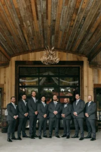 Groomsmen in gray suits with boutonnieres stand together outside the rustic barn at Whispering Pines Venue, with wooden walls and an antler chandelier in the background.