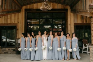 A group of bridesmaids in dusty blue gowns hold white bouquets, standing with the bride in a white lace gown outside a rustic wooden barn with an antler chandelier at Whispering Pines Venue.