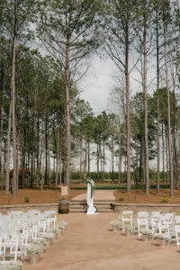 White chairs line a petal-strewn aisle in an outdoor pavilion surrounded by tall pine trees, with a floral cross and barrels at the altar for Kelsey and Andrew’s wedding ceremony.