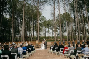 Kelsey in a white gown with a veil and Andrew in a gray suit exchange vows under a floral cross, with guests seated in white chairs and pine trees in the background.