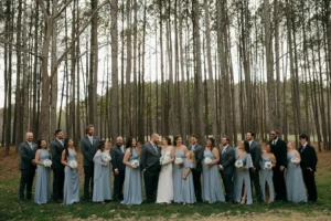 The bridal party, with bridesmaids in dusty blue gowns and groomsmen in gray suits, stands with Kelsey and Andrew in a white gown and suit, surrounded by pine trees at Whispering Pines.