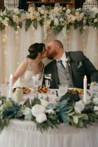 Kelsey and Andrew kiss at their sweetheart table, decorated with candles, blue flowers, and greenery, with twinkling lights and draped fabric in the background during their reception.