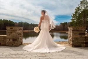 Bride Madison in a white gown with a flowing train stands by the serene lake, framed by stone pillars