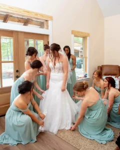 Bride in a white lace gown, is surrounded by bridesmaids in sage green dresses adjusting her veil