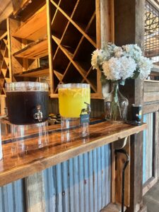 A wooden bar at Sarah’s graduation party with clear dispensers of iced tea and lemonade, accented by white hydrangeas in a glass vase, set against a rustic wooden backdrop.