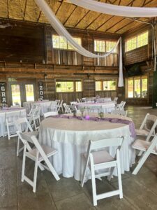 The interior of Whispering Pines Event Venue for Sarah’s graduation, featuring round tables with white linens, purple runners, and white chairs, decorated with draped fabric and string lights.