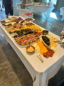 A long table at Sarah’s graduation party covered with a white cloth, displaying a charcuterie spread including cheeses, meats, fruits, pickles, dips, and crackers, with a rustic venue setting in the background.