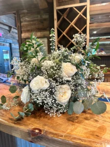 A wooden bar at Sarah’s graduation party displaying a lush centerpiece of white roses, baby’s breath, and eucalyptus, set against a rustic wooden backdrop with a wine rack design.