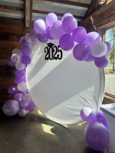 A photo backdrop at Sarah’s graduation party with a white circular backdrop, purple and white balloons, and “2025” signage with a graduation cap, set against a wooden wall with large windows.