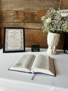 A table at Sarah’s graduation party with an open Bible, a sign asking guests to highlight their favorite verse and sign their name, a “Class of 2025” block, and a vase of white roses and baby’s breath against a wooden backdrop.