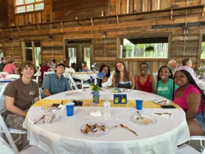 A group of guests seated around a round table at Peyton’s graduation party, enjoying a meal with yellow table runners, a floral centerpiece, and a framed photo, set against a rustic wooden interior with large windows.