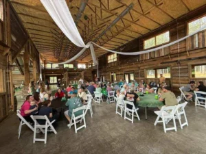 A spacious barn interior at Whispering Pines Event Venue filled with guests seated at round tables with green tablecloths, celebrating Dylan’s graduation.