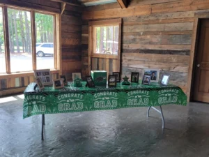A close-up of a table at Whispering Pines Event Venue decorated with a green "Congrats Grad" tablecloth, displaying framed photos, a green graduation cap, and other memorabilia, set against a rustic wooden wall with windows.