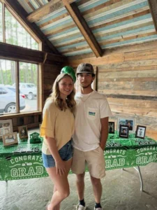 A young man and woman standing together in front of a decorated table at Whispering Pines Event Venue, covered with a green "Congrats Grad" tablecloth.