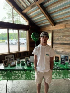 Dylan, in a white t-shirt and cap standing in front of a table at Whispering Pines Event Venue, covered with a green "Congrats Grad" tablecloth and adorned with framed photos and a balloon, with large windows and pine trees in the background.