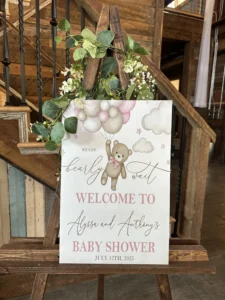 A decorative welcome sign for "Alyssa and Anthony's Baby Shower" dated July 12, 2025, featuring a teddy bear with balloons and clouds, adorned with greenery and white flowers, in Whispering Pines Venue in Henderson, NC.