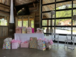 A rustic indoor setup at a baby shower with a table covered in white cloth, adorned with numerous gift bags and boxes in pink and pastel colors, featuring "baby girl" themes. Two white folding chairs are placed nearby, and large windows reveal a scenic outdoor view with trees and parked cars. A balloon with "baby" written on it hangs near the window.