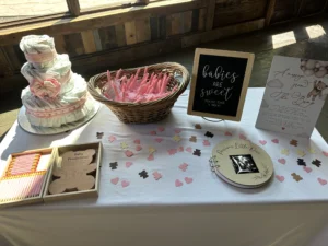 A white table decorated with a pink and white diaper cake, a basket of pink treat sticks, a wooden "Baby Predictions" box with pencils, and a "Precious Little One" guest book with an ultrasound photo. The table is sprinkled with pink and brown bear and heart confetti, with a chalkboard sign reading "babies are sweet please take a treat."