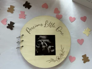 A wooden guest book with the text "Precious Little One" engraved on it, featuring a black-and-white ultrasound photo in the center, surrounded by pink and brown bear and heart confetti on a white tablecloth.