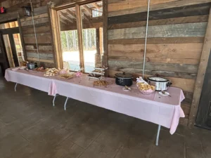 A long table covered with a pink tablecloth, set up with various foods including sandwiches, pastries, and slow cookers, in a rustic wooden interior with large windows showing greenery outside.