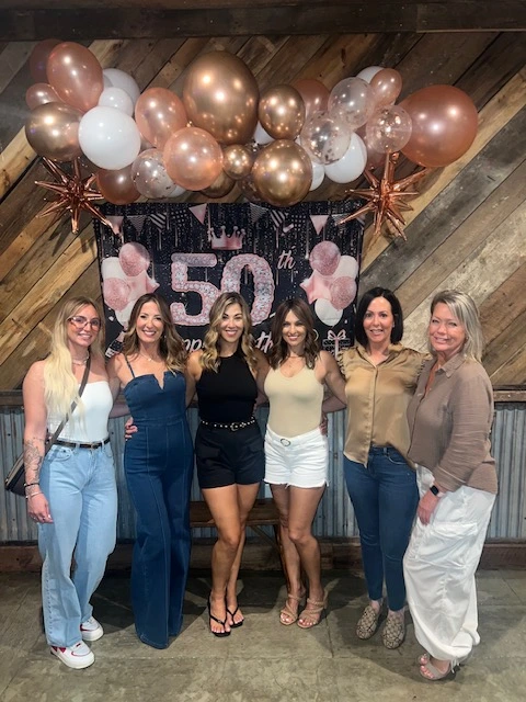 A group of friends posing in front of a "50th" banner with rose gold and white balloons at Whispering Pines Venue in Henderson, NC, celebrating a milestone birthday.