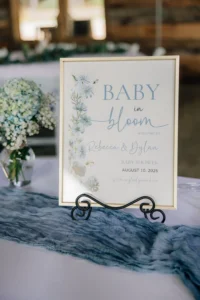A decorative sign reading 'Baby in Bloom' with the names Rebecca & Dylan and the date August 10, 2025. The sign is placed on a table with blue floral decorations and a vase of flowers at a baby shower at Whispering Pines Venue in Henderson, NC.