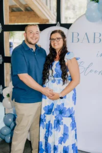 Rebecca, glowing in a blue floral maternity dress, and Dylan, in a blue shirt and khaki pants, standing together and smiling in front of a 'Baby in Bloom' backdrop with blue balloons at Whispering Pines Venue.
