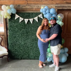 A joyful couple, Caitlyn and Connor McFalls, stand in front of a green floral wall decorated with balloons and a "Cayson" banner at their dinosaur-themed baby shower at Whispering Pines Venue. Caitlyn, pregnant, wears a blue striped dress, while Connor stands beside her in a black shirt and shorts. The rustic venue features wooden walls, a vintage chair, and colorful balloons in blue, green, and yellow.