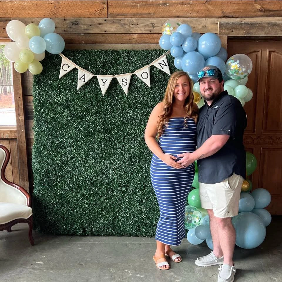 A joyful couple, Caitlyn and Connor McFalls, stand in front of a green floral wall decorated with balloons and a "Cayson" banner at their dinosaur-themed baby shower at Whispering Pines Venue. Caitlyn, pregnant, wears a blue striped dress, while Connor stands beside her in a black shirt and shorts. The rustic venue features wooden walls, a vintage chair, and colorful balloons in blue, green, and yellow.