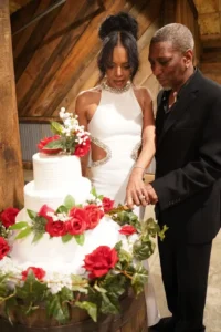 Derrick and Elethia smiling together as they cut their anniversary cake at Whispering Pines Venue, surrounded by elegant red and white floral décor.