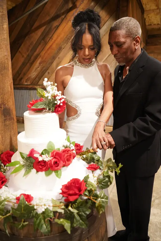 Derrick and Elethia smiling together as they cut their anniversary cake at Whispering Pines Venue, surrounded by elegant red and white floral décor.