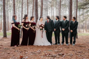 Bridal party posing outdoors beneath tall winter pine trees at Whispering Pines Venue, capturing a whimsical woodland wedding aesthetic with elegant seasonal attire.
