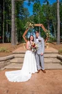 Newlyweds Andrew and Taylor pose beneath the floral ceremony arbor surrounded by towering pine trees at Whispering Pines Venue in Henderson, NC.