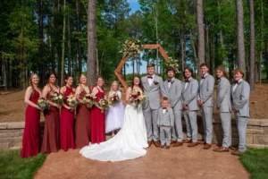 Bride, groom, bridesmaids, groomsmen, and flower girl gather for a wedding party portrait in front of the outdoor ceremony site at Whispering Pines Venue.