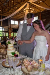 Andrew and Taylor smile as they cut their elegant two-tier wedding cake during their rustic reception, surrounded by warm lighting and wood accents at Whispering Pines Venue.
