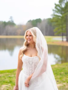Bride Zoey Elliott smiling during her bridal portrait session beside the pond at Whispering Pines Venue in Henderson, NC, wearing a lace wedding gown and flowing veil.