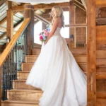 Bride Zoey Elliott poses on the wooden staircase inside the rustic barn at Whispering Pines Venue in Henderson, NC, holding a colorful bridal bouquet.