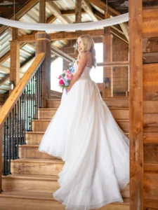 Bride Zoey Elliott poses on the wooden staircase inside the rustic barn at Whispering Pines Venue in Henderson, NC, holding a colorful bridal bouquet.