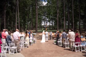Bride and groom exchanging vows during an outdoor wedding ceremony surrounded by family, friends, and pine trees at Whispering Pines Venue in Henderson, NC.