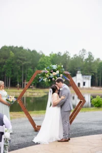 Bride and groom share a kiss beneath a floral hexagon arbor overlooking the pond at Whispering Pines Venue in Henderson, NC.