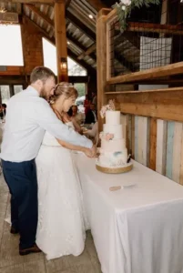 Bride and groom cut their elegant tiered wedding cake together inside the rustic barn at Whispering Pines Venue.