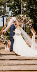 Bride and groom share a kiss beneath a floral-adorned wooden arbor during their outdoor wedding ceremony at Whispering Pines Venue.