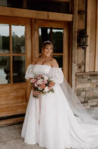 Bride in an elegant white wedding gown poses with a blush and peach floral bouquet outside the barn at Whispering Pines Venue.
